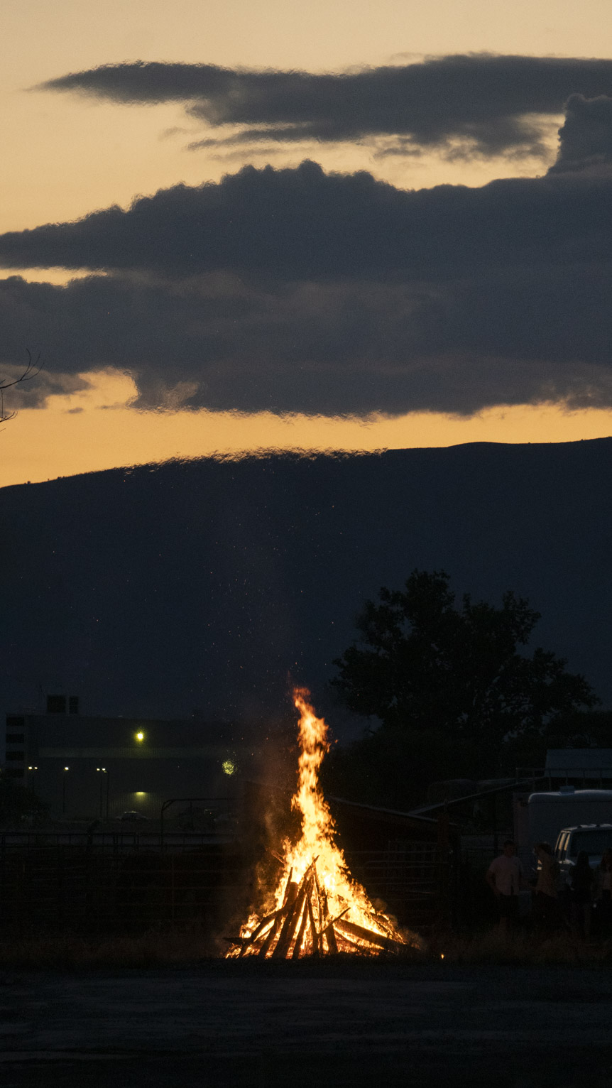 A bonfire bright at dusk whose heat hazes the mountains and clouds above